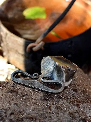 Traditioneller Feuerstahl aus geschmiedetem Eisen, rustikal gestaltet, präsentiert mit einem Feuerstein auf einem Stein und einem Topf  im Hintergrund